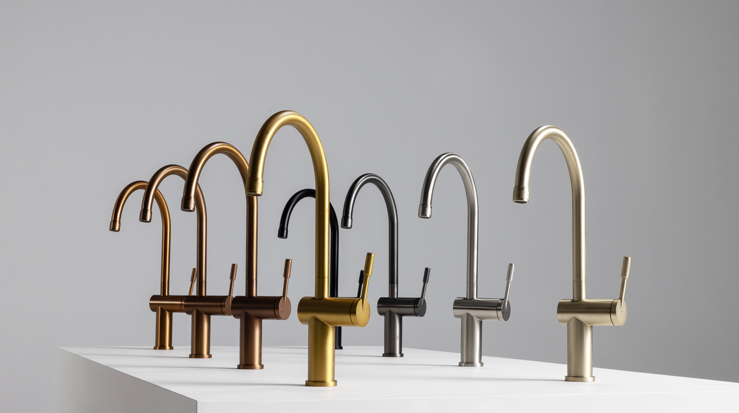 Image of Titan curved gooseneck kitchen mixer taps lined up in a grid against a gray background, displaying the full color range including gold, aged brass, brass, gunmetal, matte black, stainless steel, and rose gold, showcasing a variety of stylish kitchen tap options.