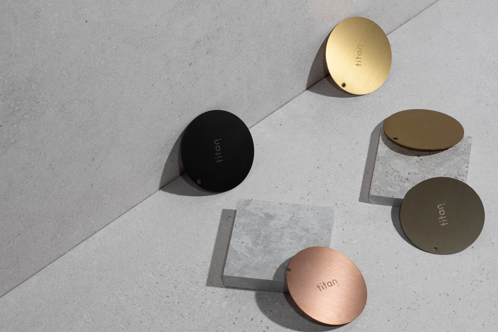 Still life photo of Titan finish samples in matte black, royal gold, rose gold, and pearl gold, arranged on a gray background with complementary gray set props, showcasing the variety of available finish samples.
